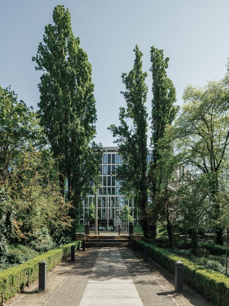 Der Garten des Hamburger Bahnhofs in vollem Grün, vom Gebäudeeingang zum Tor hin blickend. Hohe Bäume flankieren den Eingang.