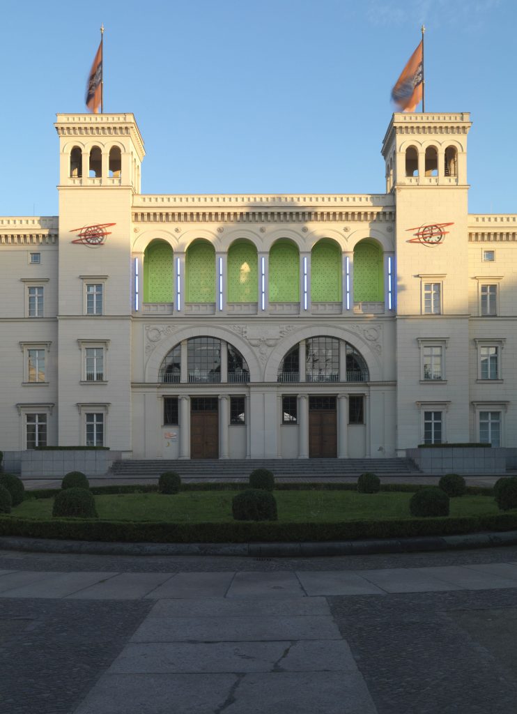 Symmetrische Fassade des historischen Bahnhofsgebäudes des Hamburger Bahnhofs mit zwei Türmen: Auf dem östlichen Turm liegt eine rote Zickzack-Linie aus Metall über der Turmuhr, auf dem westlichen Turm eine entsprechende rote Zickzack-Linie über der Schmuckrosette, die Arbeit von Ayşe Erkmen.